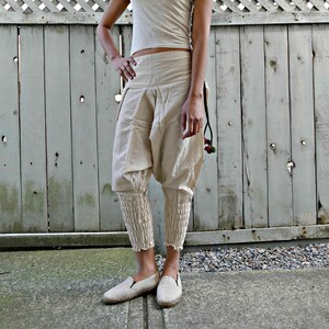 Op de afbeelding: Een vrouw draagt een wit tanktop en een beige broek met elastische manchetten. De broek is getailleerd en heeft een losse, vloeiende pasvorm. Ze staat voor een houten hek.