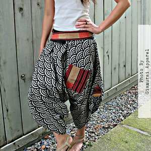 May include: A woman wearing a white tank top and black and white patterned pants with a red and black fabric pocket. She is wearing a brown and red woven belt. The pants have a wide leg and are gathered at the waist. The woman is standing on a gravel path.