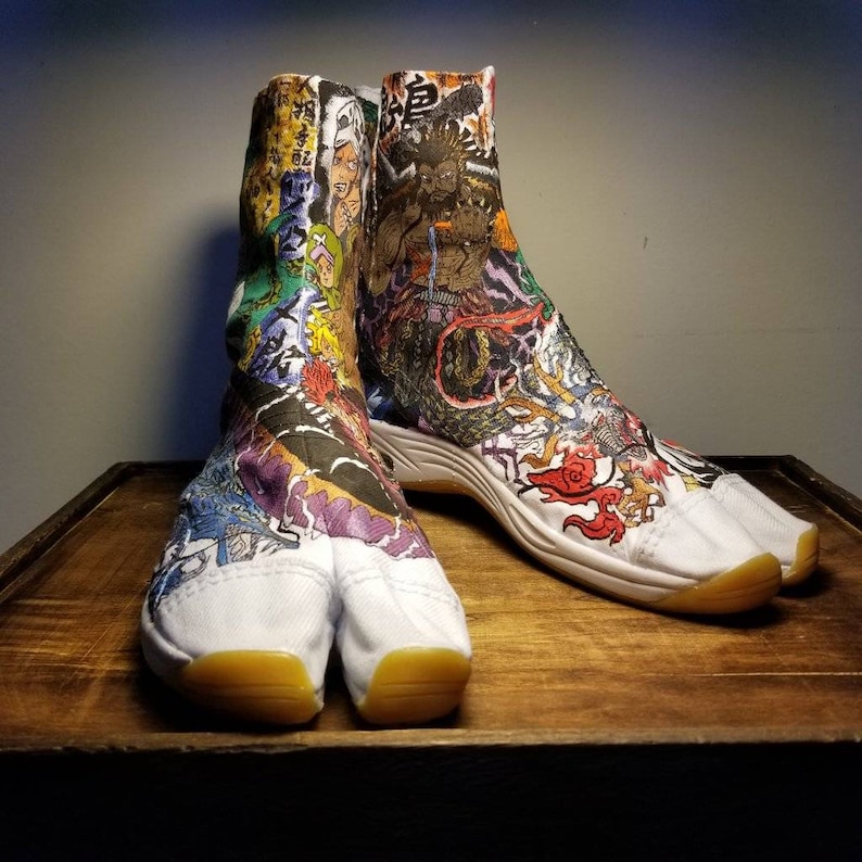 May include: A pair of white tabi shoes with a split toe design. The shoes are decorated with colorful Japanese-inspired artwork, including dragons, flowers, and characters. The shoes are sitting on a wooden surface.
