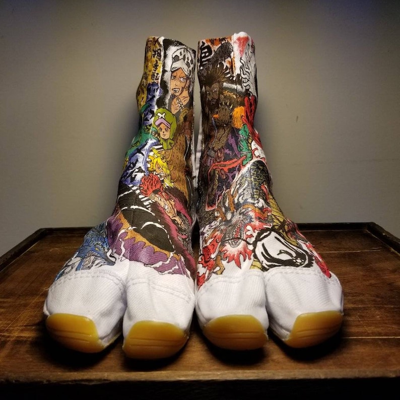 May include: A pair of white tabi shoes with a colorful cartoon design. The shoes have a split toe and a yellow sole.