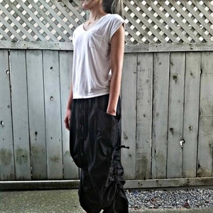 May include: A woman wearing a white t-shirt and black pants with a drawstring detail. She is standing in front of a wooden fence.