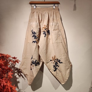 May include: Beige pants with a floral print in shades of blue and brown. The pants have a wide leg and a drawstring waist.