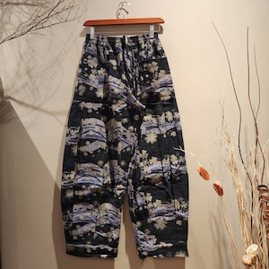 May include: Black patterned pants with a floral and wave design. The pants are hanging on a wooden hanger. The fabric has a dark background with light-colored floral and wave patterns.