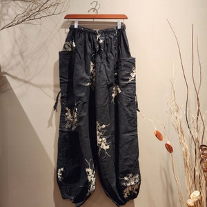 May include: Black trousers with a white floral pattern and drawstring ties. The trousers have two large pockets on the sides.