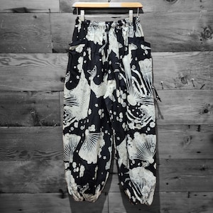 May include: Black and white patterned pants with a koi fish and fan design. The pants are hanging on a wooden hanger against a wooden background. The pants have pockets and a drawstring waist.