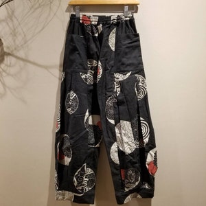 May include: Black pants with a white, red, and black circular pattern. The pants have an elastic waistband, pockets, and a loose fit. The fabric appears to be a lightweight cotton or linen blend. The pants are hanging on a wooden hanger.