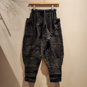 May include: Dark grey, loose-fitting trousers with a drawstring waist and large side pockets. The trousers are hanging on a wooden hanger against a neutral-coloured wall. The fabric has a subtle pattern.