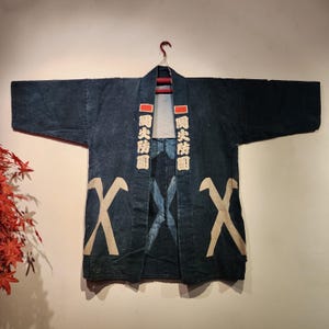 May include: A dark blue Japanese fireman's coat, hanging on a red hanger. The coat features white Japanese characters and stylized white 'X' designs. The coat is made of a textured fabric and has a faded appearance.