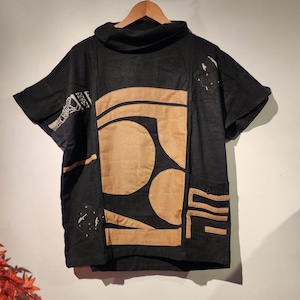 May include: Black short-sleeved shirt with a high collar and tan geometric shapes. The shirt features a unique design with tan fabric patches and a pocket on the lower right. It is displayed on a wooden hanger.