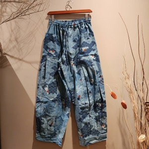 May include: Light blue patterned pants with a drawstring waist, featuring a repeating wave and crane design in navy blue and red. The pants are hanging on a wooden hanger, showcasing a relaxed fit and comfortable style.