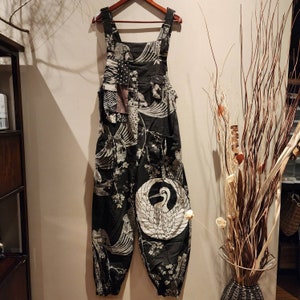 May include: Black and white patterned overalls featuring a crane design on the leg. The overalls have adjustable straps, pockets, and a unique patchwork detail on the chest. The fabric is adorned with a Japanese-inspired print.