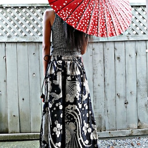 May include: A woman wearing a black and white patterned skirt with a red and white umbrella over her head. The skirt has a floral and fish design. The woman is standing in front of a wooden fence.