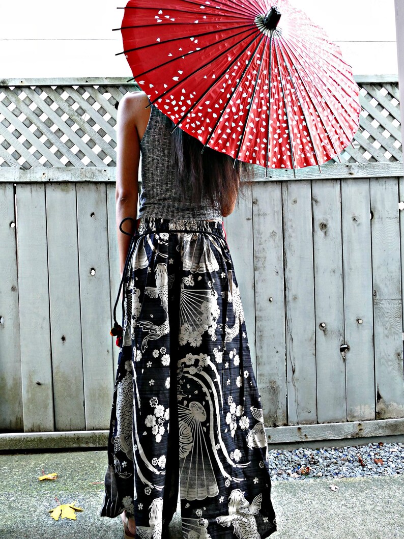 May include: A woman wearing a black and white patterned skirt with a red and white umbrella over her head. The skirt has a floral and fish design. The woman is standing in front of a wooden fence.