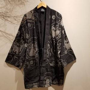 Puede incluir: Bata negra estilo kimono con un estampado gris y blanco con diseños circulares y figuras. La bata tiene un corte holgado, mangas largas y dos bolsillos delanteros. Cuelga de una percha de madera sobre un fondo neutro.