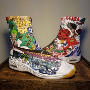 May include: A pair of white tabi shoes with a colorful anime design. The shoes feature characters from the anime One Piece, including Luffy, Zoro, and Sanji. The shoes are decorated with Japanese characters and symbols.