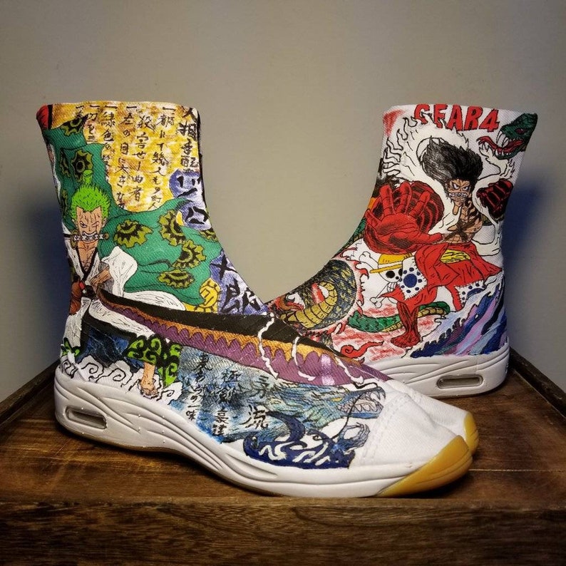 May include: A pair of white tabi shoes with a colorful anime design. The shoes feature characters from the anime One Piece, including Luffy, Zoro, and Sanji. The shoes are decorated with Japanese characters and symbols.