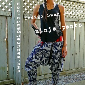 May include: A woman wearing a black tank top with the text "Indigo Swan Design" and "ninja pants" printed on it. She is wearing blue and white patterned pants with a red sash. She is holding a black sword in her right hand and is standing in front of a wooden fence.