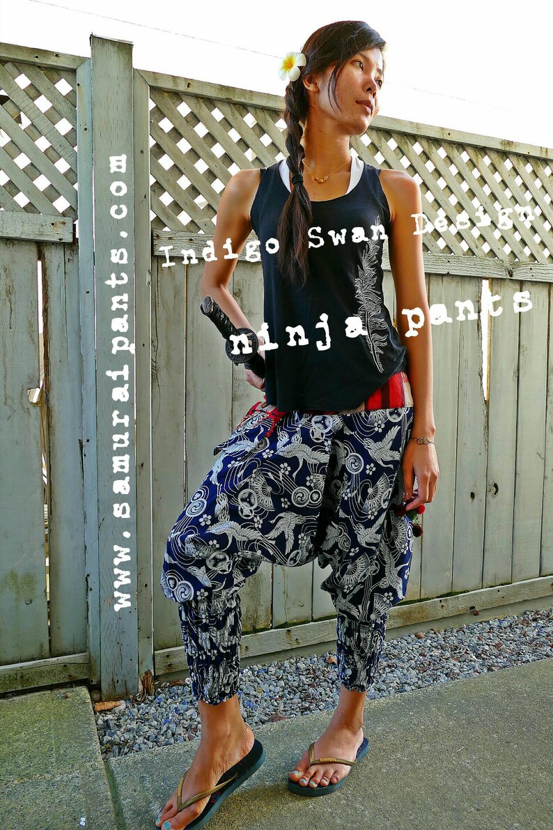 May include: A woman wearing a black tank top with the text "Indigo Swan Design" and "ninja pants" printed on it. She is wearing blue and white patterned pants with a red sash. She is holding a black sword in her right hand and is standing in front of a wooden fence.
