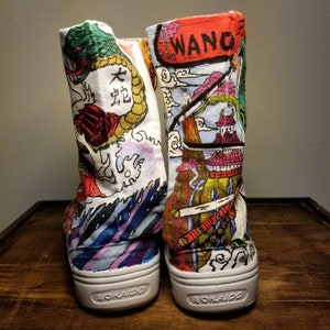 May include: A pair of white canvas high-top shoes with colorful hand-painted designs. One shoe features a dragon and waves, the other features a Japanese castle and the word "WANO". Both shoes have the word "SOKAIDO" printed on the soles.
