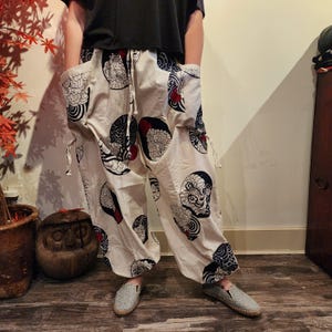 May include: Cream-colored pants with a repeating pattern of black and navy blue circular designs, accented with red details. The pants have large pockets and a drawstring waist. The person is wearing a black top and gray slip-on shoes.