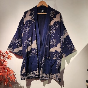 May include: A navy blue kimono-style robe with a crane and floral pattern. The robe has two front pockets and a wave pattern along the bottom edge. The cranes are beige, and the flowers are white.