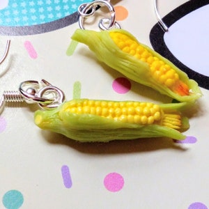May include: A pair of silver earrings with corn on the cob charms. The corn is yellow and green and has a realistic look.