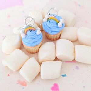 Pode incluir: Um par de brincos de cupcake azul com um arco-íris e nuvens brancas no topo. Os brincos são exibidos em uma superfície branca com marshmallows brancos.