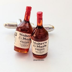 May include: Cufflinks featuring miniature Maker's Mark whiskey bottles with red wax seals and white labels. The bottles are brown with the text "Maker's Mark Whisky". The cufflinks have a silver-colored metal backing.