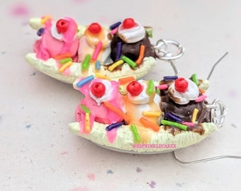 Puede incluir: Un par de pendientes caprichosos de banana split. Cada pendiente presenta un barco de plátano en miniatura con bolas de helado rosa, vainilla y chocolate, cubierto con una cereza y chispas. Los pendientes están hechos con un metal de color plateado.
