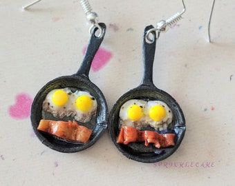 Puede incluir: Un par de pendientes con miniaturas de sartenes de hierro fundido con huevos fritos y tocino. Las sartenes son negras y los huevos son amarillos con yemas blancas. El tocino es marrón y crujiente.