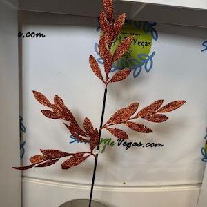 May include: A decorative artificial branch featuring multiple pointed leaves covered in a vibrant orange glitter finish. The branch is supported by a thin black stem that is anchored into a round metal container filled with a light-colored foam base.