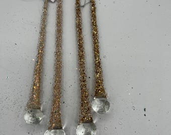 Könnte beinhalten: Vier goldfarbene, mit Perlen besetzte Weihnachtsschmuckstücke mit klaren, tropfenförmigen Böden. Jedes Ornament hat eine Schlaufe zum Aufhängen und ein Etikett mit der Aufschrift "MELROSE". Die Ornamente liegen auf einer weißen Oberfläche mit verstreutem Glitzer.