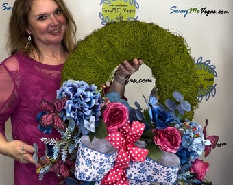 May include: A green moss wreath decorated with blue and pink flowers, butterflies, and a blue and white ribbon with pink polka dots. The wreath is held by a person wearing a pink shirt.