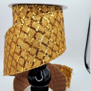 May include: Gold ribbon with a geometric pattern and sequins. The ribbon is wrapped around a black stand.