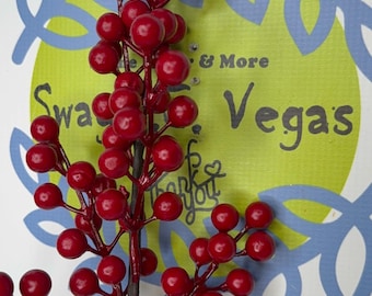 Könnte beinhalten: Ein dekorativer künstlicher Zweig mit zahlreichen glänzenden, hellroten Beeren an einem dunkelbraunen Stiel. Der Zweig ist vor einem kreisförmigen Logo mit hellgrüner Mitte, blauen Blattumrissen und dem Text Swags & More Vegas platziert.