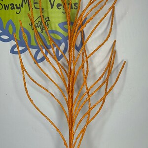 May include: Orange glittered decorative branch. The artificial stem has multiple thin branches and is displayed in a white vase. The background includes the text "SwayMeVegas.com" and "Home Deco & More".