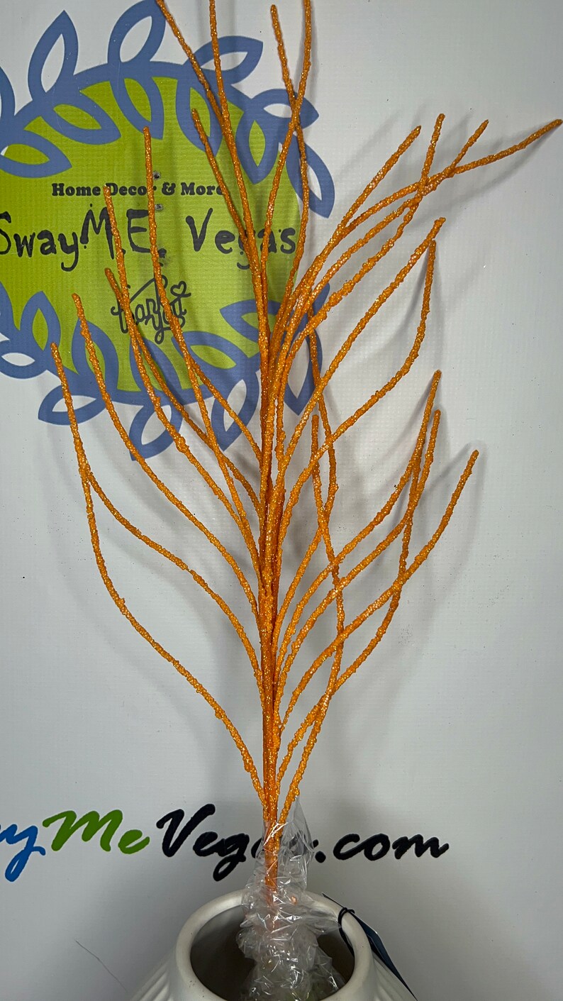 May include: Orange glittered decorative branch. The artificial stem has multiple thin branches and is displayed in a white vase. The background includes the text "SwayMeVegas.com" and "Home Deco & More".