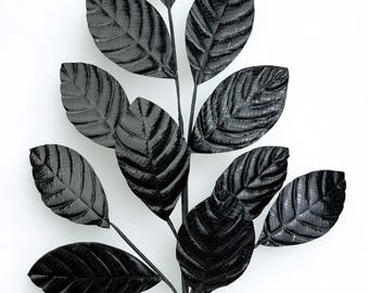 May include: A black artificial plant stem with multiple textured leaves. The leaves have a detailed veining pattern and are arranged along a dark stem. The plant is set against a white background.