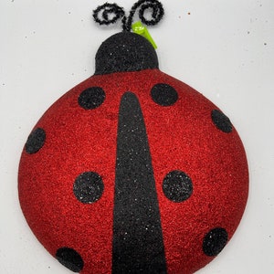 May include: Red ladybug with black spots and a black stripe down the middle. The ladybug has black antennae and is made of glitter.