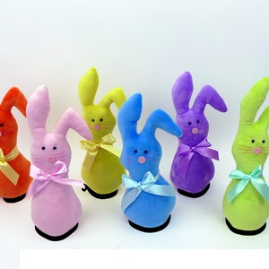 May include: Six plush Easter bunny toys in different colors: orange, pink, yellow, blue, purple, and green. Each bunny has a bow tied around its neck.