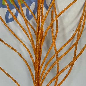 May include: Close-up of an orange glittered decorative branch. The branch has multiple stems with a textured, sparkling surface. The background is a light color with some text visible. This decorative branch is suitable for floral arrangements and seasonal decor.