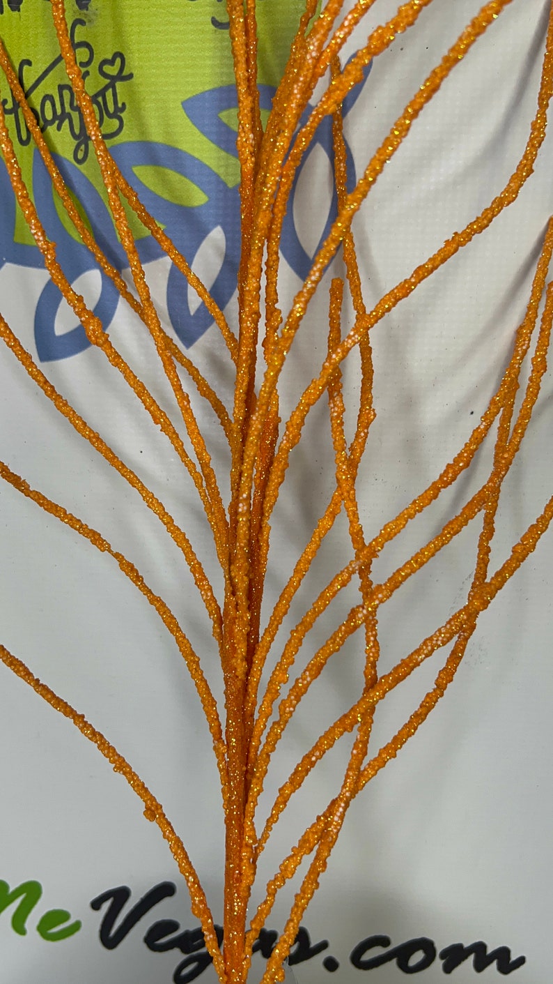 May include: Close-up of an orange glittered decorative branch. The branch has multiple stems with a textured, sparkling surface. The background is a light color with some text visible. This decorative branch is suitable for floral arrangements and seasonal decor.