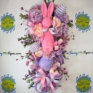 May include: A decorative Easter wreath featuring a pink bunny, faux flowers, and decorative eggs. The wreath is adorned with pink and purple ribbons and is designed for door or wall decor. The text "Sway Me Vegas.com" is visible.