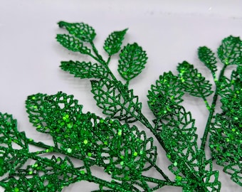 Könnte beinhalten: Grüne Glitzerblätter auf weißem Hintergrund. Die Blätter sind aus einem dünnen, zarten Material gefertigt und haben eine glitzernde Oberfläche.