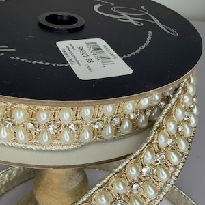 May include: A spool of decorative ribbon with a gold metallic weave, adorned with faux pearls and clear rhinestones. The ribbon is on a wooden spool, with a black top. The label reads "PENDELOQUE" and "made in India".
