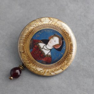 May include: A round, gold-framed pin with a portrait of a woman in a red and white dress. The pin has a dark red glass bead attached to the bottom.