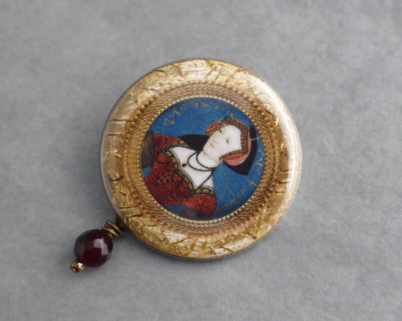 May include: A round, gold-framed pin with a portrait of a woman in a red and white dress. The pin has a dark red glass bead attached to the bottom.