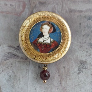 May include: A gold circular pendant with a portrait of a woman in a red and white dress. The pendant is surrounded by a gold frame and has a red bead hanging from it.