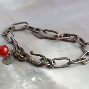 May include: A dark brown metal bracelet with a red bead and a small metal charm. The bracelet is made of linked oval shapes.