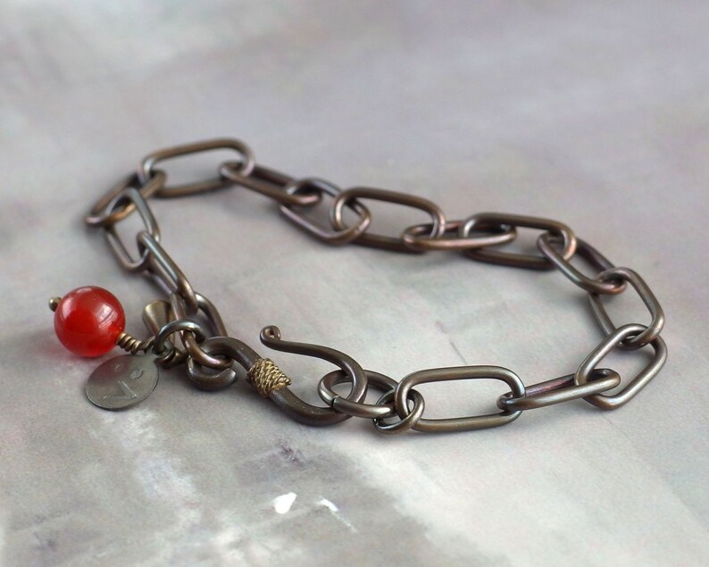 May include: A dark brown metal bracelet with a red bead and a small metal charm. The bracelet is made of linked oval shapes.
